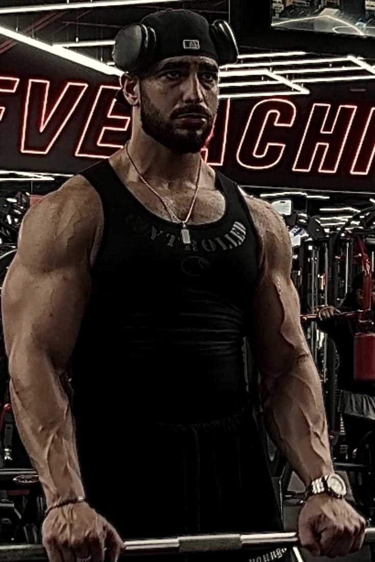 Hadeed in a black tank top, holding a curl bar in the gym